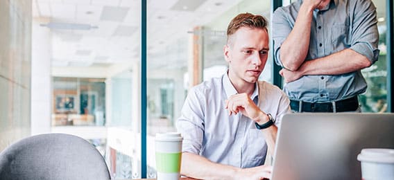 karriere tutor Zwei Männer in einer modernen Büroumgebung. Einer sitzt und arbeitet an einem Laptop, neben ihm steht eine Kaffeetasse, der andere steht mit der Hand am Kinn daneben. Beide sind auf den Laptop-Bildschirm konzentriert und diskutieren eine Online-Weiterbildung.