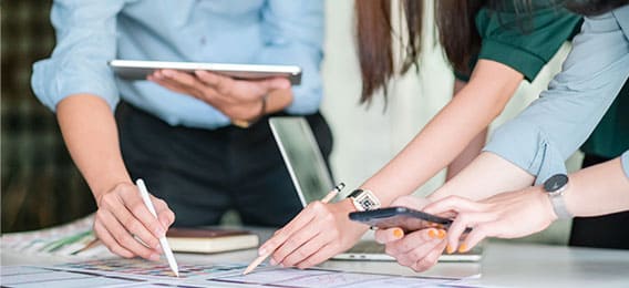 karriere tutor Drei Personen arbeiten an einem Tisch, der mit Dokumenten und Diagrammen bedeckt ist. Zwei Personen verwenden Stifte, um auf die Papiere zu zeigen, während die dritte Person ein Smartphone hält. Die Umgebung scheint ein Büro oder ein Arbeitsplatz zu sein, der Weiterbildungsmöglichkeiten durch die Agentur für Arbeit anbietet.